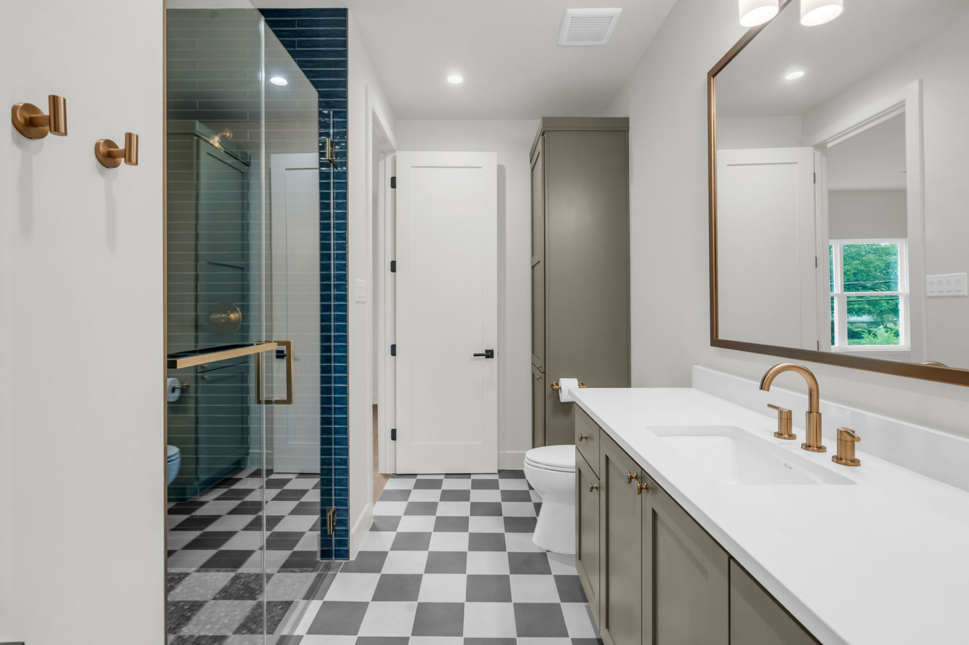 Guest bath with a glass shower framed in deep blue tile, sage cabinetry, brass fittings and checkerboard flooring.