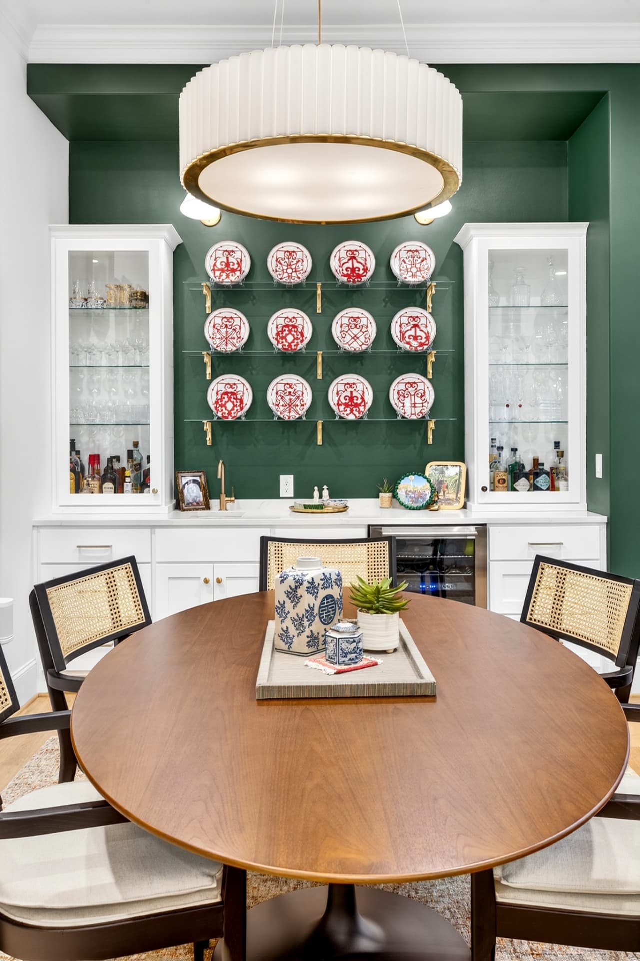 Dining room with deep green walls, a plate display on glass shelves, white built-in bar cabinetry and a round walnut table.
