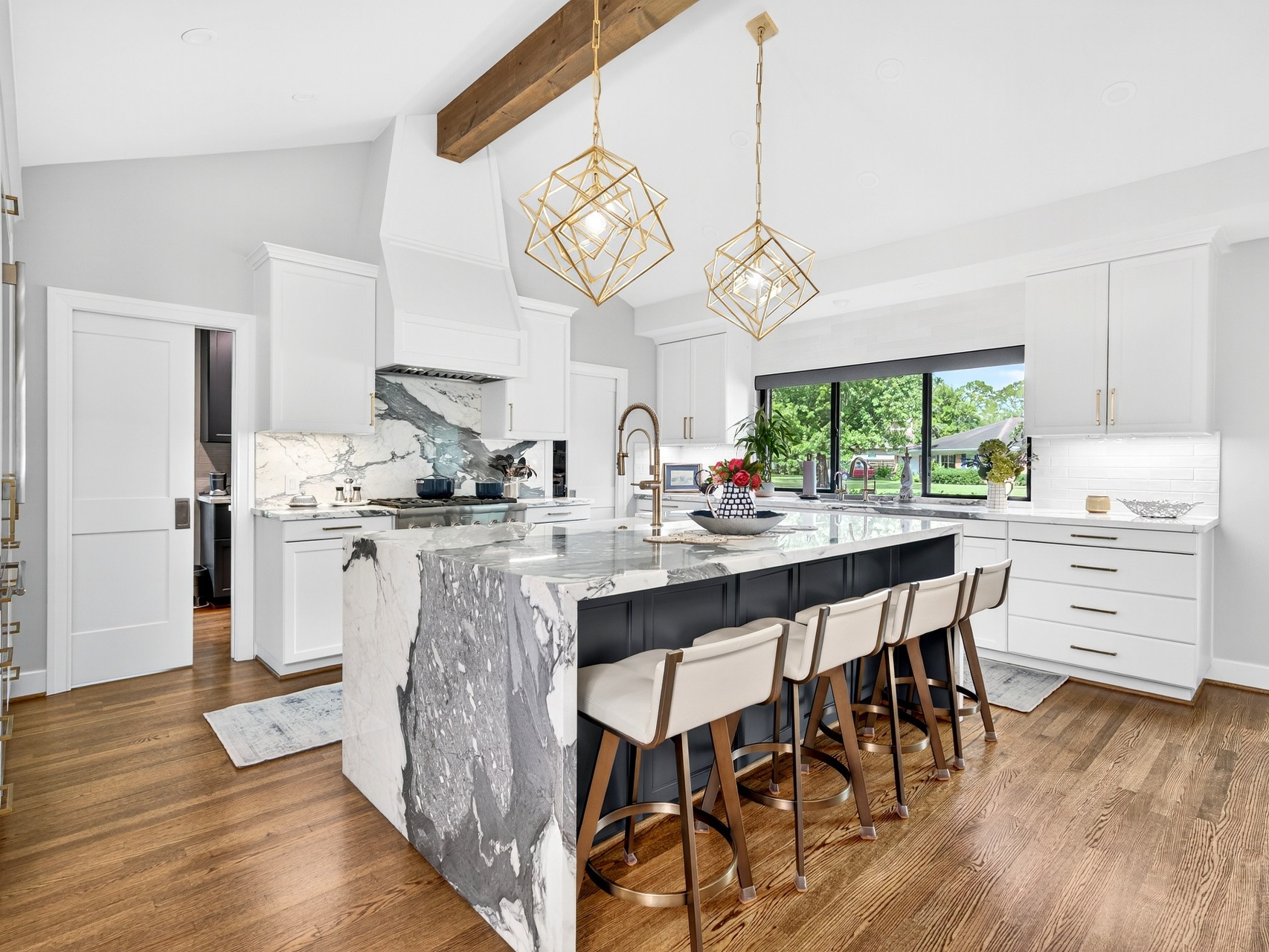 A renovated kitchen with a marble waterfall island, brass pendants, white cabinetry, and warm wood floors.