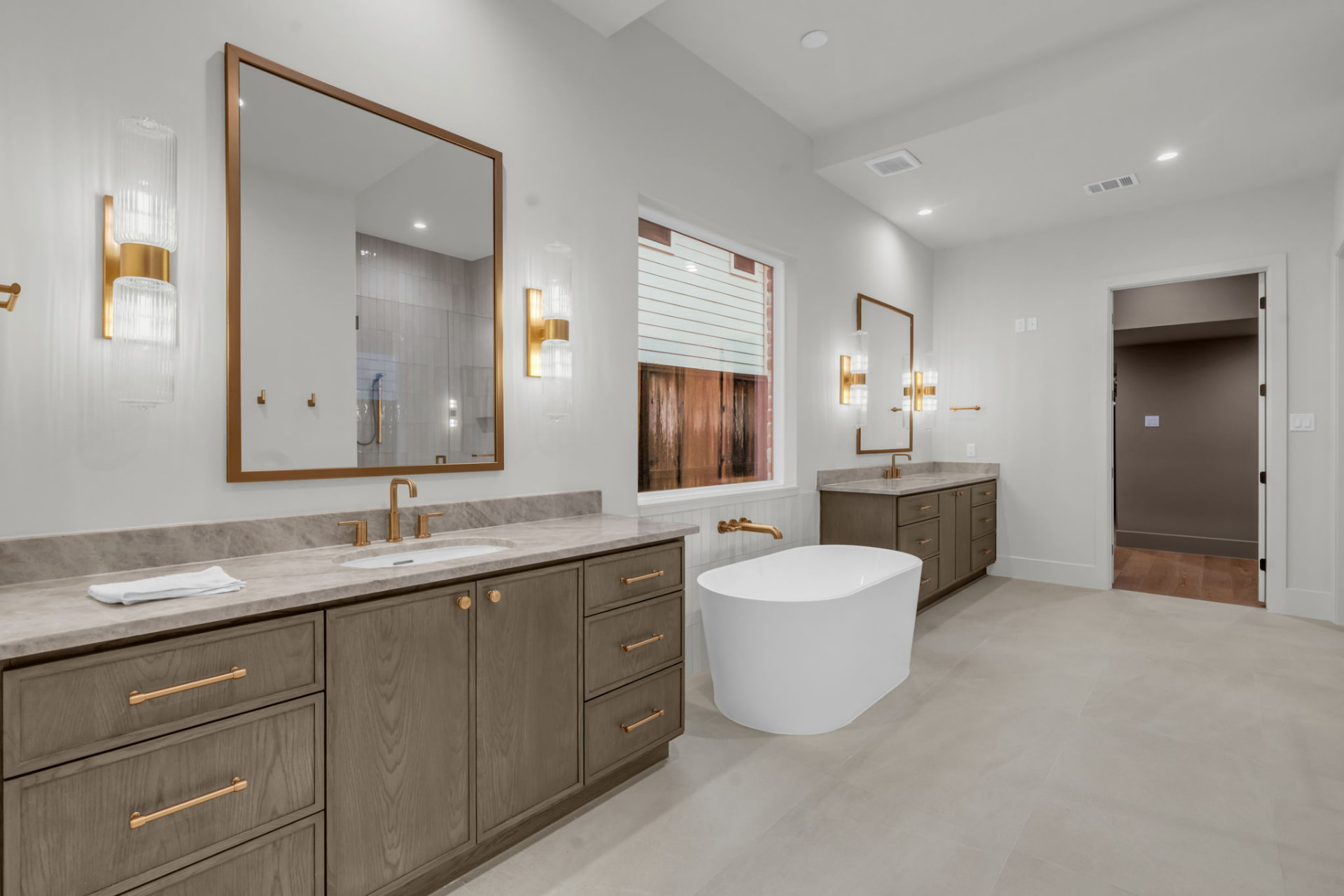 Primary bath with paired oak vanities, marble counters, brass sconces and a freestanding soaking tub beneath a window.