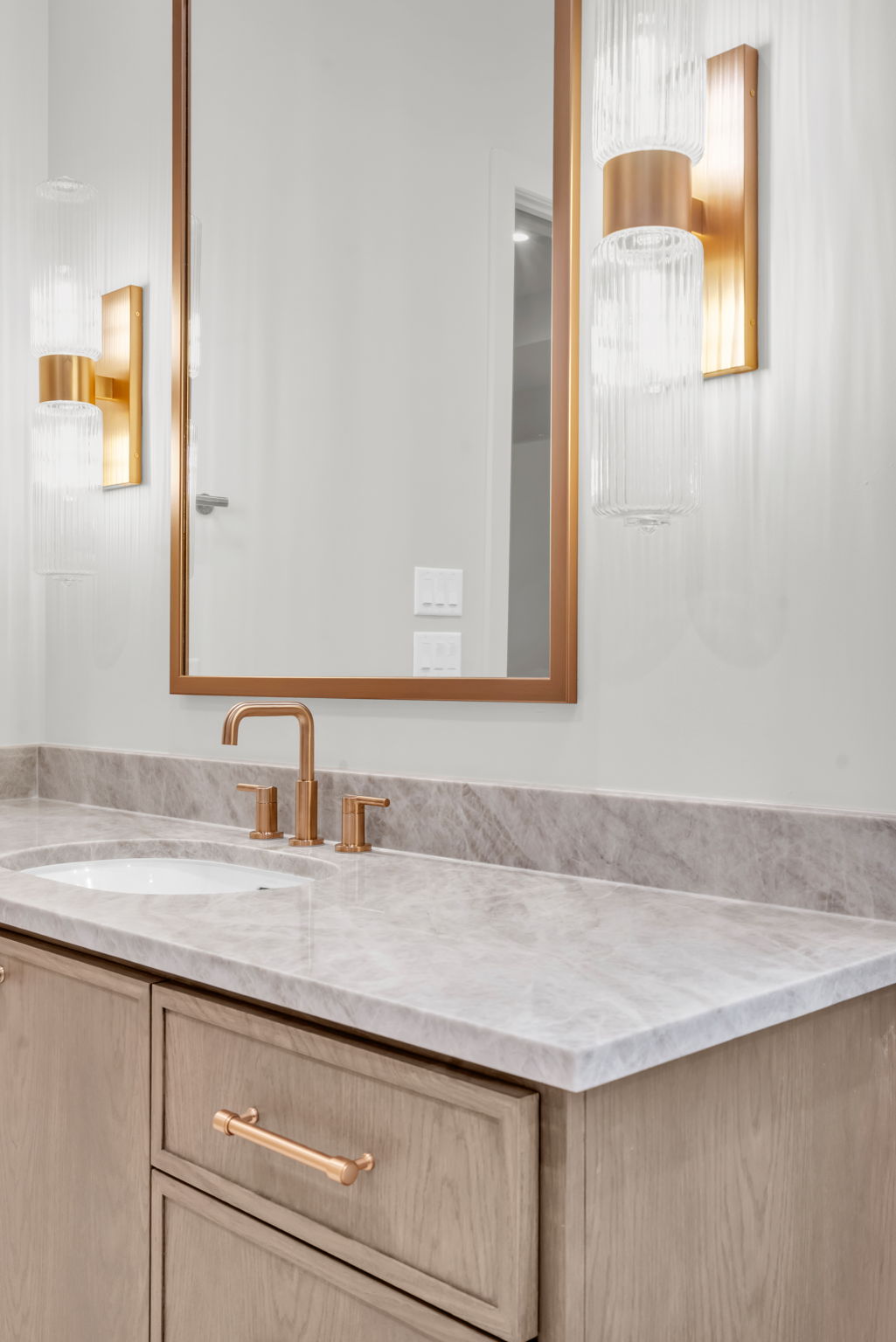 Powder vanity in soft oak with a honed marble countertop, brass faucet and fluted glass sconces flanking a brass-framed mirror.