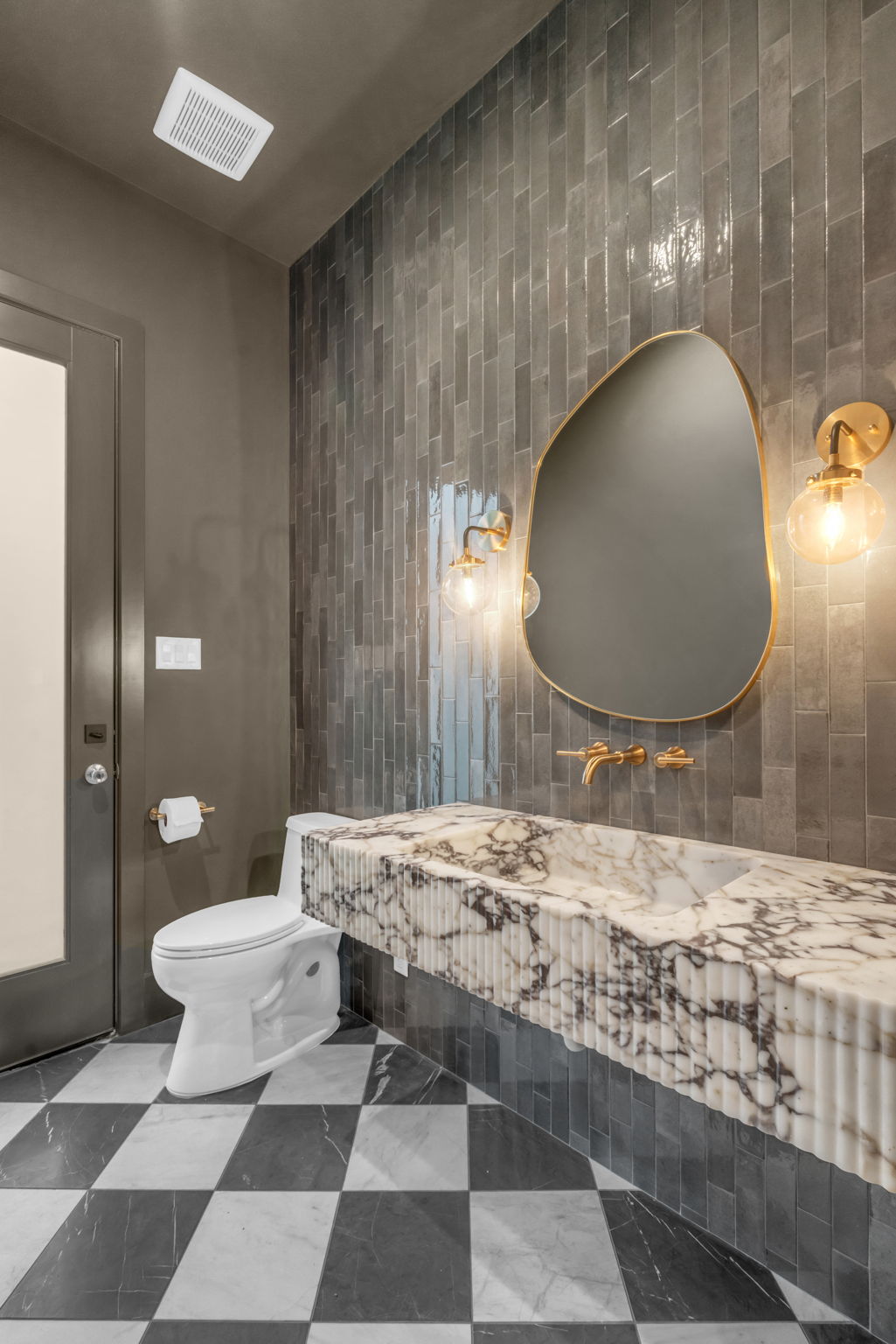 Powder room with glazed charcoal tile, a fluted marble trough sink, organic brass mirror and checkerboard marble floor.